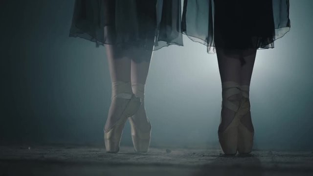 Female feet in pointe standing in pose in spotlight on a black background. Two girls simultaneously stand on their toes. Professional ballerinas dancing in ballet shoes in studio. Slow motion.