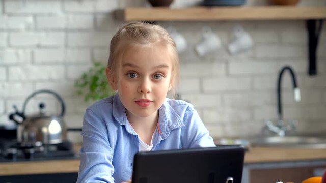 Cute little girl looking at the tablet at the kitchen. Smiling, looking at the camera and wave hand.