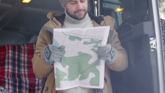 Tilt up of male tourist in warm clothes standing near his car with map in his hands and examining it