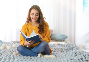 Young beautiful woman in warm sweater reading book on bed at home, space for text
