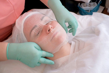 A young girl relaxes in a beauty salon. Beautician covers gauze woman's face. Preparation for...