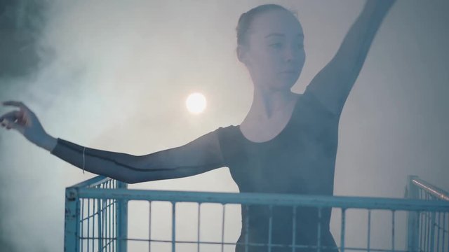 Close up portrait of graceful professional ballerina dancing in black dress in the studio inside the blue cage in spotlight on a black background. Young beautiful woman raising hand standing in a