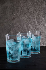 Three full glasses of soda-based cocktail and blue Curacao liqueur with ice stand in a row on a dark textured background. Concept: skull in a glass of ice cubes, hidden threat, danger, dosage.