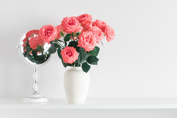 Roses flowers of coral color in vase and mirror on shelf against white wall. Space for text