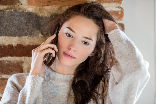 Young brunette woman talking on smartphone while standing against brick wall