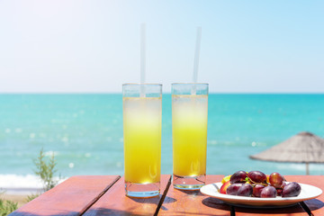 Two glasses of fresh tropical fruit juice on the beach, and a plate with grapes and watermelon on the table, against the sea. Holiday and vacation concept