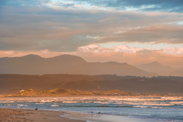Fototapeta na wymiar Rays of golden evening light falling on the Plettenberg Bay beach at sunset, with mountains in the distance. Garden Route, Western Cape, South Africa