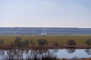 Picturesque landscape: river, green field, mountain and cloudless blue sky. Smoke from the fire in the center