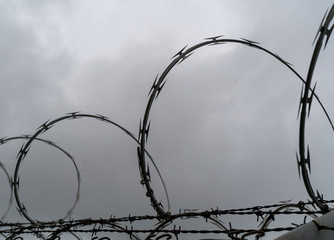 Razor wire isolated and silhouetted at the top of a barbed-wire fence with dark clouds in background