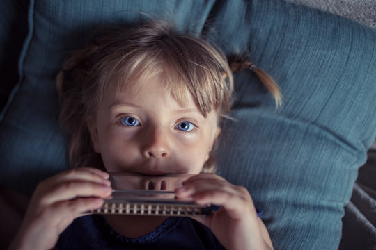 Portrait of blond girl with gray eyes playing harmonica while lying on sofa at home