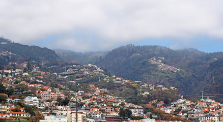 a wide scenic panoramic aerial cityscape of the city of funchal in Madeira with buildings of the city in front of mountains with the tops covered by white clouds