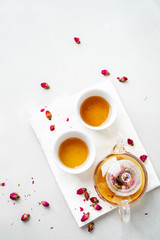 Top view of two white cups and transparent teapot with herbal tea of dried pink roses buds over textile napkin on gray background with copy space. Brewing and Drinking tea.