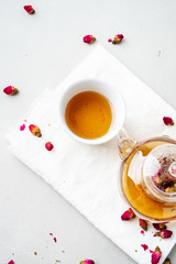 Top view of two white cups and transparent teapot with herbal tea of dried pink roses buds over textile napkin on gray background with copy space. Brewing and Drinking tea.