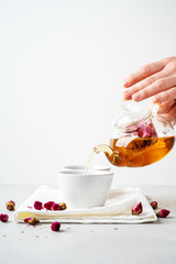 Female hands pouring tea of dry pink rose buds from transparent kettle to teacup on white background with copy space. Brewing and Drinking tea.