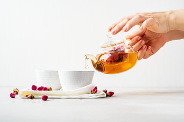 Female hands pouring tea of dry pink rose buds from transparent kettle to teacup on white...