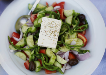 Delicious traditional Greek salad with tomatoes, cucumbers, pepper, olives, green salad, spices and feta cheese served on a white plate.