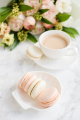 Elegant sweet dessert macarons, cup of coffee and pastel colored beige flowers bouquet on white marble