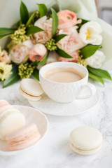 Elegant sweet dessert macarons, cup of coffee and pastel colored beige flowers bouquet on white marble