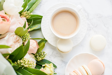 Elegant sweet dessert macarons, cup of coffee and pastel colored beige flowers bouquet on white marble
