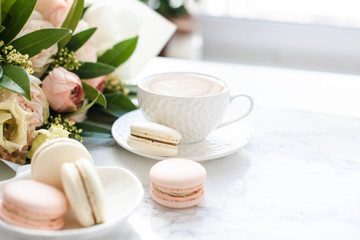 Elegant sweet dessert macarons, cup of coffee and pastel colored beige flowers bouquet on white marble