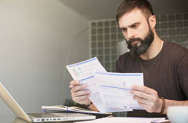 Bearded young man sitting at table looking at documents and thinking. Business man going through paperwork at home office.