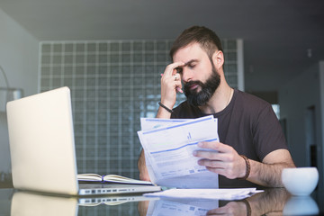 Bearded serious concentrated man working at laptop and sitting at the table while looking at the papers and holding his head with hand.