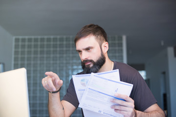 Bearded serious concentrated man working at laptop and sitting at the table while looking at the...