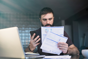 Bearded young man working with laptop at home browsing bills and documents . Businessman going through paperwork at home office.