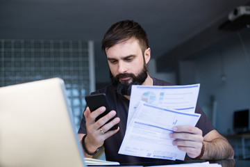 Bearded young man working with laptop at home browsing bills and documents . Businessman going through paperwork at home office.