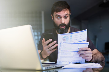 Bearded positive man working with laptop at home browsing documents . Businessman going through paperwork at home office with unpaind bills.