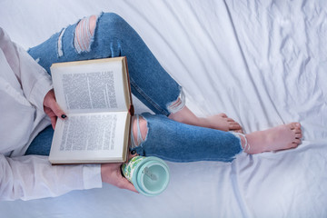 Girl student in blue jeans on leg and bare feet lying in cozy bed and reading book and holding retro coffee cup. Relaxation and education concept. Close up, selective focus