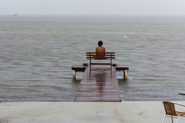 A man sits on a bench in front of a lake during a rainy day.