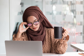 Young asian muslim woman sitting alone in a cafe working on her laptop and holding a coffee mug.