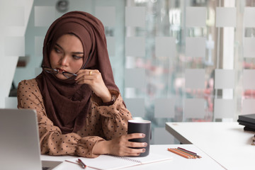 Young asian muslim woman sitting alone in a cafe working on her laptop and holding a coffee mug.