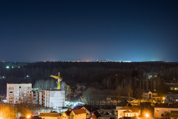 The outskirts of the night city. Private houses and the construction of a new apartment building. Lanterns on the street with cottages. Crane on the background of the forest.
