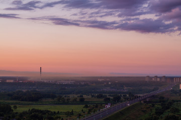 District of the city at dusk. Smoky plume from the enterprise. Release of dirty air into the environment. Satellite City on the horizon in the evening. Road on the outskirts of the town at sunset.