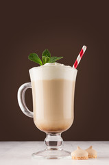 Irish coffee with cream, fresh mint and striped red straw in transparent glass with handle on white wood board in brown color background, vertical.