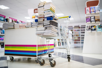 Shopping cart full books in bookstore shop near the shelves with books.