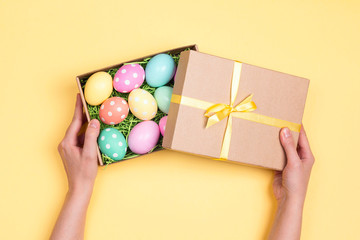 Female hands hold gift box with easter eggs on yellow background.