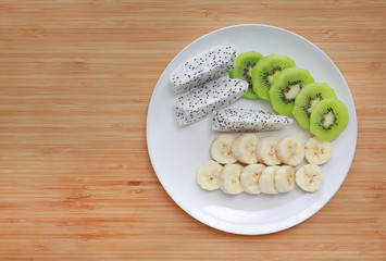 Fresh sliced fruit (banana, dragon fruit and kiwi) on white plate against wooden board background with copy space.