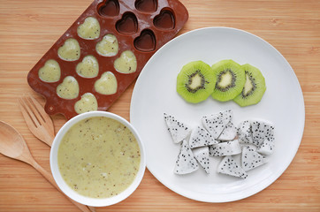 Baby food mashed in container on wooden board for freezing with plate of sliced dragon fruit and kiwi.