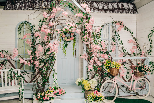 the interior of a rustic photo studio, the facade of a white building with a porch decorated with blooming flowers, baskets with flowers next to the steps and a pink bicycle
