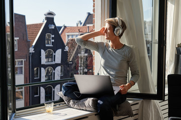 Woman with short hair sitting near opened window at sill with canal view with laptop and listening to music in headphones. Relaxed happy mood.