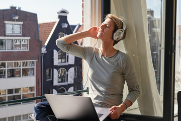 Woman with short hair sitting near opened window at sill with canal view with laptop and listening to music in headphones. Relaxed happy mood.