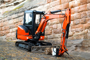 Small machine res excavator on sidewalk during repair works on street in urban environment - obrazy, fototapety, plakaty