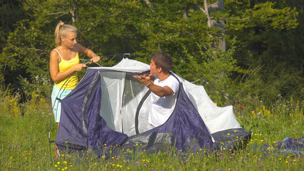 Annoyed girlfriend argues with boyfriend while they set up their tent by forest.