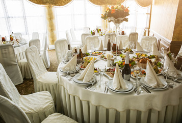 Beautiful white round table at the restaurant. Wedding reception day. Champagne and floral composition in vase. Tasty food.