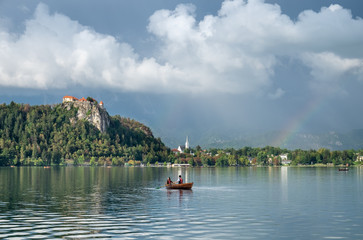 Fototapeta na wymiar Amazing photo of Lake Bled at evening after rain with vibrant rainbow on the sky and couple in wooden boat. Slovenia