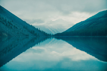 Naklejka na ściany i meble Amazing silhouettes of mountains and low clouds reflected on mountain lake. Beautiful ripples on water mirror. Cloudy sky in highlands. Atmospheric ghostly landscape. Wonderful mystic mountainscape.