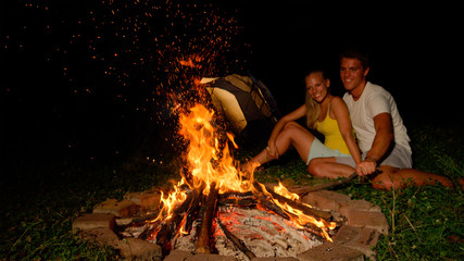 CLOSE UP: Cheerful man strikes campfire with stick while snuggling with girl.